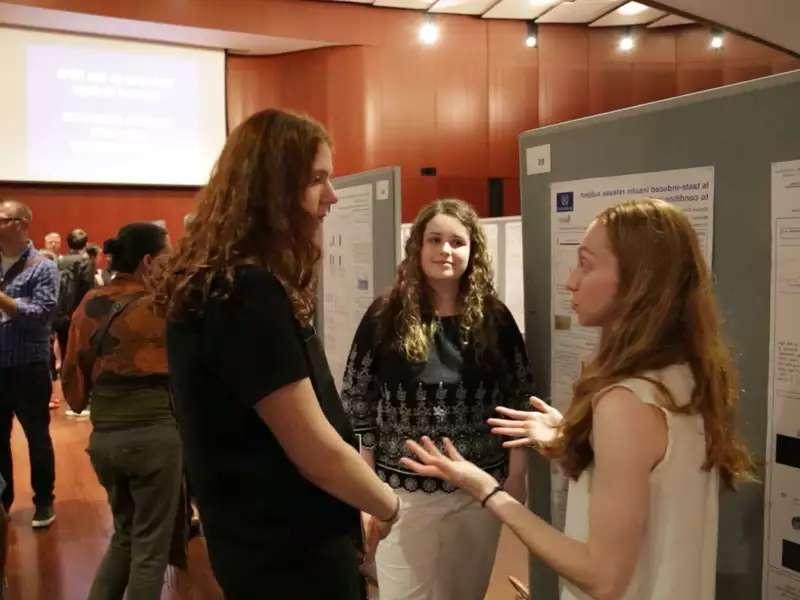 3 white women students talking in front of a large poster 